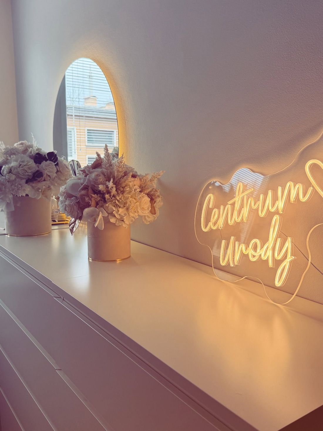 Neon Centrum Urody, kwiaty i podświetlane lustro na białej komodzie